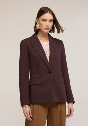 Vrouw die een donkerbruine blazer draagt over een kanten top, beige broek, grote gouden oorbellen en gouden armbanden, staand tegen een effen achtergrond.