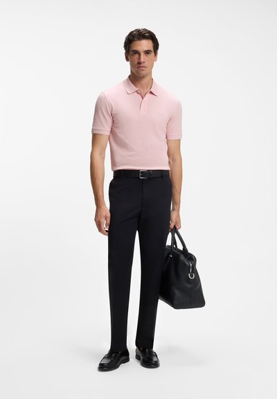 Homme portant un polo rose, un pantalon noir, une ceinture noire et des chaussures noires, tenant un sac à main en cuir noir dans sa main droite.