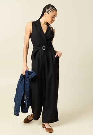 Femme aux cheveux tressés portant une combinaison noire sans manches ceinturée et des chaussures marron, tenant une veste en jean bleu dans une main.