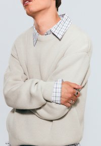Suéter de punto beige con un ajuste holgado, que presenta un acabado texturizado. Debajo, una camisa blanca con un patrón de cuadros negros, visible en el cuello.