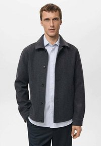 Giacca di lana grigio scuro con collo a risvolto, chiusura frontale con bottoni e maniche lunghe. Indossata sopra una camicia azzurra a righe con bottoni.