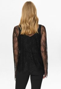 Must mustrikkidega must pitsbluus, millel on pikad varrukad, V-kaelus ja läbipaistev muster. Alumine äär on kergelt kõver, musta aluskihi peal.