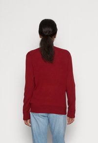 Červený svetr s kulatým výstřihem, s žebrovanými manžetami a lemem, vyrobený z měkké látky. Nositelný s světle modrými džínami s volným střihem. Zobrazen pohled zezadu.