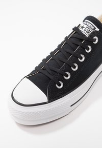 Schwarze Leinenschuhe mit weißer Gummi-Spitze und -Sohle, ausgestattet mit schwarzen Schnürsenkeln, silbernen Ösen und einem Logotag auf der Zunge.