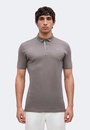 Uomo con capelli scuri e ricci che indossa una polo grigia aderente con bottoni e pantaloni bianchi, in piedi davanti a uno sfondo chiaro e uniforme.