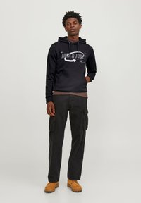 Sudadera con capucha negra con estampado de logo gris, combinada con pantalones cargo negros con bolsillos laterales y botas de trabajo marrones.