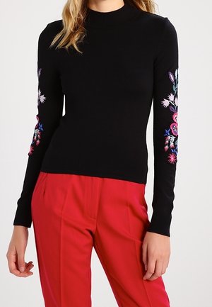 Kvinde iført en tætsiddende sort sweater med farverig blomsterbroderi på ærmerne og højtaljede, knaldrøde bukser mod en hvid baggrund.