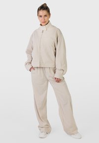 Kobieta w beżowej kurtce na zamek i pasujących szerokich spodniach, stojąca pewnie z rękami w kieszeniach na jednolitym tle.