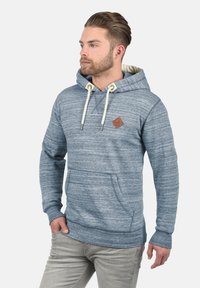 Sudadera azul jaspeada con un bolsillo canguro, cordones y una pequeña etiqueta de cuero marrón. Textura de tela suave y corte casual.