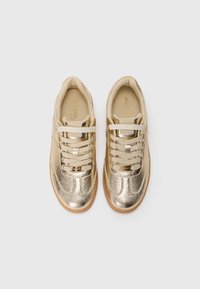 Un paio di sneakers basse metalliche dorate con lacci beige e suole in gomma, viste dall'alto su uno sfondo semplice.