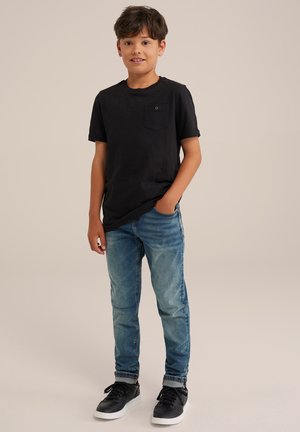 T-shirt nera a maniche corte con taschino sul petto, abbinata a jeans in denim blu chiaro e sneakers nere con suole bianche.