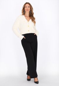 Vrouw met lang golvend haar in een witte donzige vest met knopen, zwarte wijde broek, zwarte hakken en lange hangende oorbellen, staand met haar handen in haar zakken.