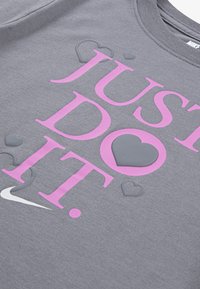 Sudadera gris con "JUST DO IT" en letras rosas negritas. Presenta acentos de corazón de tamaños variados. Logo de Nike en blanco en la parte inferior.