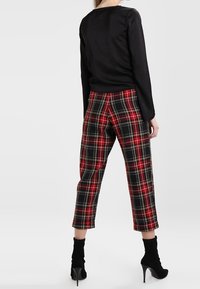 Femme portant un haut noir à manches longues, un pantalon court à carreaux rouges et noirs, et des bottines à talons hauts noires, debout de dos.