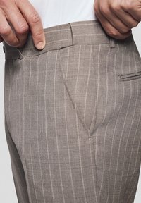 Beżowe spodnie w paski wykonane z teksturowanej tkaniny, z płaskim paskiem w talii, bocznymi kieszeniami i subtelnymi prążkami jako detal projektowy.