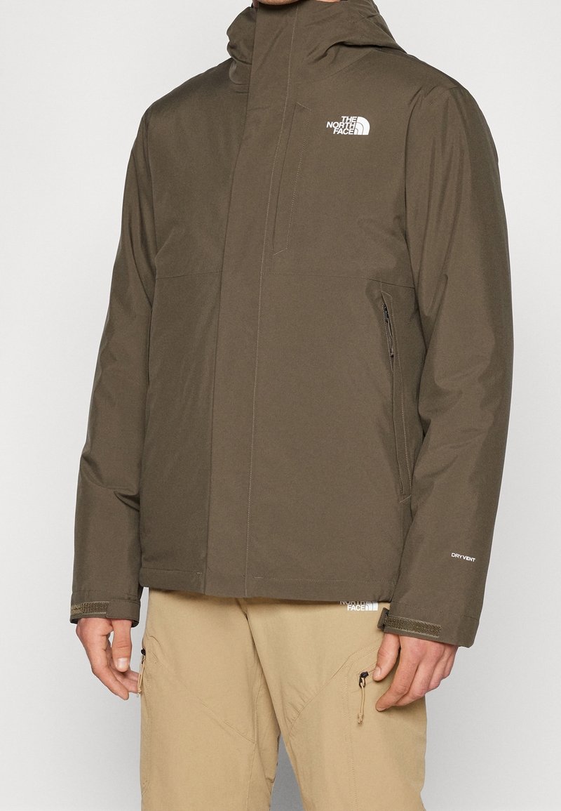 Personne portant une veste imperméable vert olive avec capuche et un pantalon cargo beige, debout devant un fond uni.