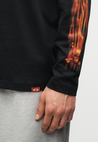 Μαύρο πουκάμισο με μακριά μανίκια και πορτοκαλί σχέδιο φλόγας στο μανίκι, κόκκινη ετικέτα μάρκας κοντά στο τελείωμα, φορεμένο με ανοιχτό γκρι παντελόνι, με ένα χαλαρό χέρι να φαίνεται.