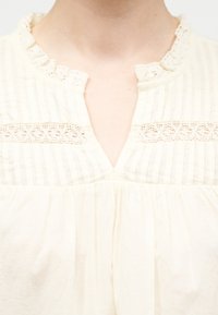 Κοντινό πλάνο κρεμ μπλούζας με δαντελένιο τελείωμα, γιακά με βολάν και κάθετες πιέτες γύρω από το λαιμό και την περιοχή του στήθους.