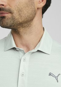 Polo gris clair en tissu texturé, avec deux boutons au col et un logo brodé foncé sur la poitrine gauche.