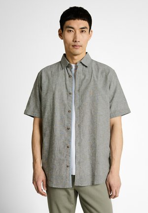 Aziatische man met een grijs overhemd met korte mouwen over een wit T-shirt en olijfgroene broek, staand tegen een witte achtergrond.