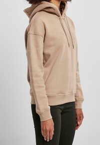 Sudadera con capucha beige, con cordones, puños y dobladillo acanalados, textura suave, corte relajado, mostrada en una persona contra un fondo liso.