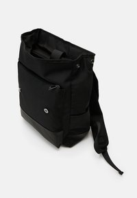 Sac cabas en tissu noir à texture lisse, doté d'une poche avant avec fermeture éclair et boutons-pression, d'une sangle en filet réglable, et d'un bas contrasté.