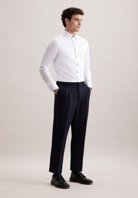 Witte button-up shirt met een smalle kraag, gecombineerd met hooggesneden marineblauwe broek. Model draagt zwarte schoenen. Eenvoudig, strakke ontwerp.