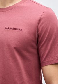 Κοντινό πλάνο ενός ατόμου που φοράει ροζ σκόνη κοντομάνικη μπλούζα με κεντημένο το λογότυπο "Peak Performance" στο αριστερό στήθος.