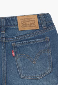Blå denimjeans med en brun läder baklapp, med ett broderat hjärtdesign på bakfickan och en röd etikett. Klassisk 5-fickors stil.