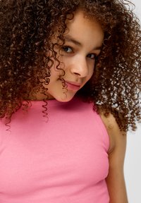 Roze geribbelde mouwloze top met een ronde hals, gecombineerd met krullend haar tot op de schouders, benadrukt door een rijke, natuurlijke textuur en glans.