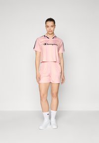 Roosa lühike sporditopp ja sobivad lühikesed püksid, mis on valmistatud siledast ja kergest materjalist; mustade aktsentidega ja "Champion" logo ees.