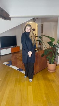 Mulher de cabelo comprido vestindo um casaco escuro, calças pretas e sapatos prateados, em pé numa sala de estar próximo a um sofá castanho e uma grande planta em vaso.