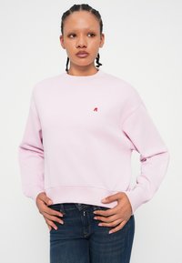 Ροζ φόρμα φτιαγμένη από μαλακό ύφασμα, με στρογγυλή λαιμόκοψη και ριμπ μανσέτες. Ένα μικρό κόκκινο λογότυπο είναι εμφανές στο στήθος.