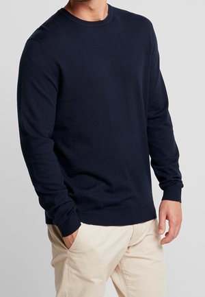 Man wearing a navy blue long-sleeve crewneck sweater and beige pants with one hand in pocket against a plain background.