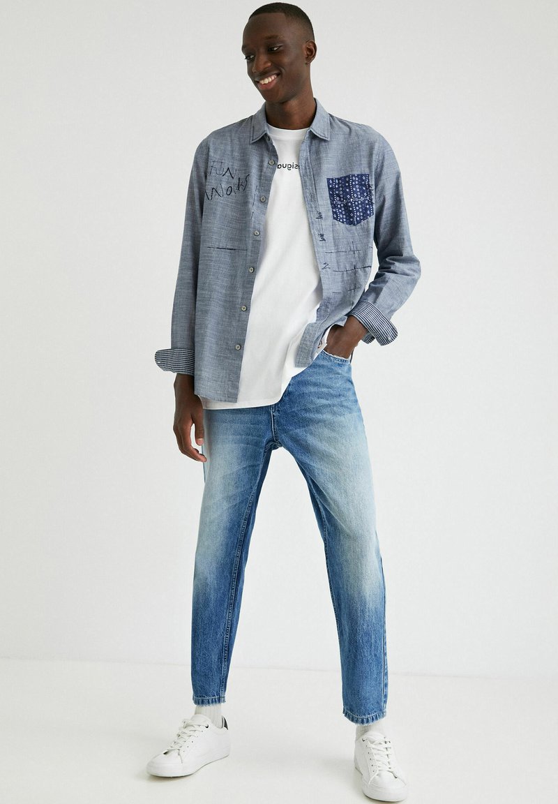 Hellblaues Baumwollhemd mit blauem gemustertem Taschen. Kombiniert mit einem weißen T-Shirt und hellblauen, schmal geschnittenen Jeans. Weiße Sneakers.