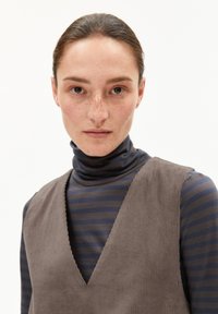 Brauner Cordwesten mit tiefem V-Ausschnitt, getragen über einem navy- und grau gestreiften Rollkragenpullover. Der Stoff wirkt strukturiert. Glatte Frisur.
