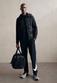 Sudadera negra con cremallera y patrón texturizado, camiseta negra de cuello redondo, pantalones negros con un logo blanco de Nike y zapatillas deportivas grises.