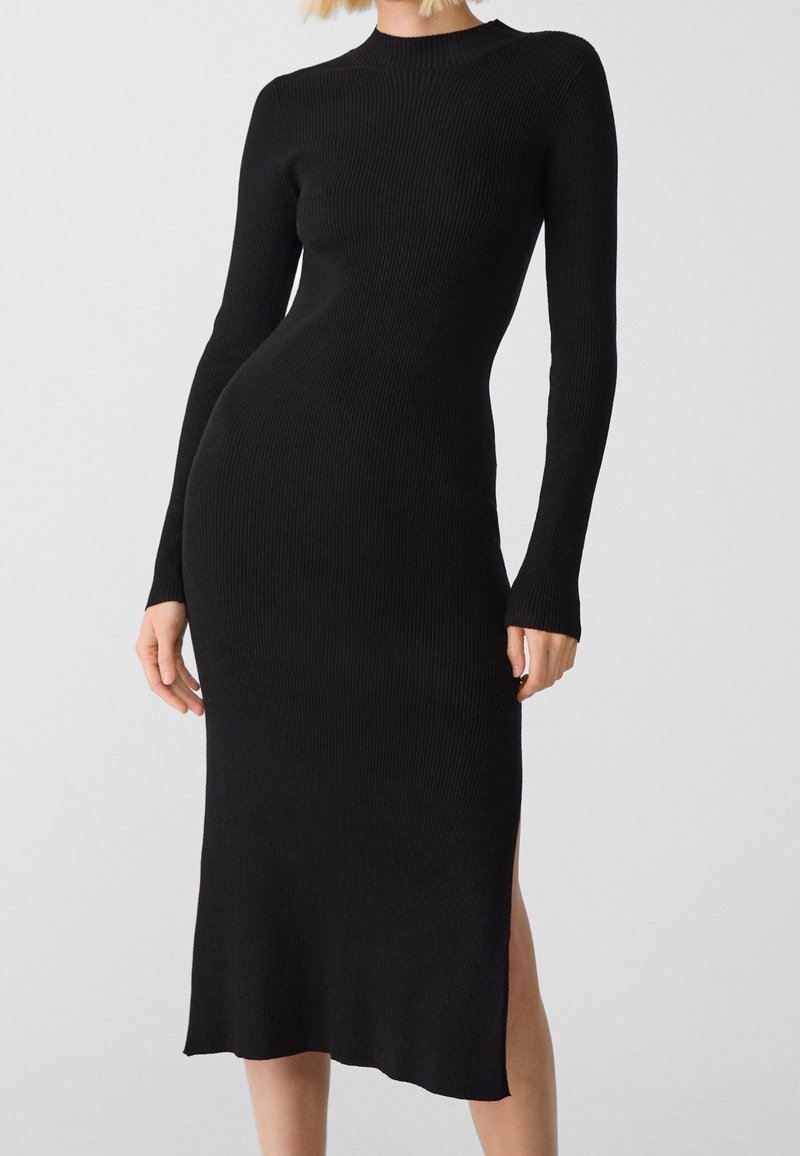 Robe midi ajustée en maille côtelée noire à manches longues, col montant et fente latérale, portée par une personne debout sur un fond uni.