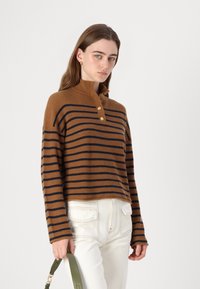 Pull marron côtelé avec des rayures bleu marine, col montant et trois boutons dorés. Assorti à un pantalon blanc taille haute et un sac vert.