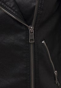 Giacca in pelle nera con zip metalliche, finitura testurizzata e numerose tasche con cerniera. Primo piano di dettagli di zip e cuciture.