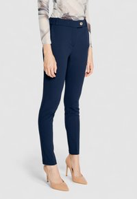 Pantalons slim bleu marine avec une texture lisse, dotés d'une fermeture à bouton sur le devant, de poches latérales et de coutures ajustées. Chaussures à talons nus.
