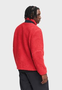 Czerwona polarowa kurtka z wysokim kołnierzem, teksturowaną powierzchnią i długimi rękawami. Krój jest luźny, co zapewnia swobodny fason.