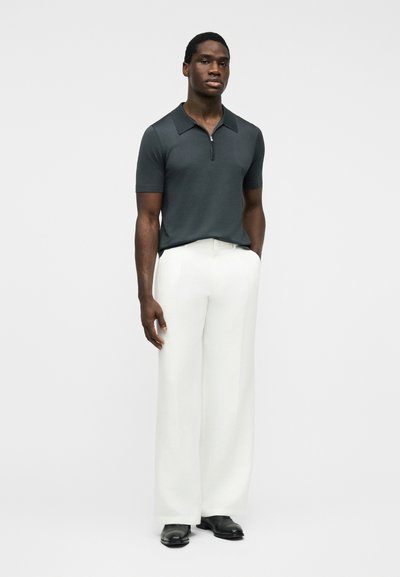 Homme portant une chemise zippée à manches courtes foncée, un pantalon blanc à jambes larges et des chaussures noires, debout contre un fond clair uni avec une main dans la poche.