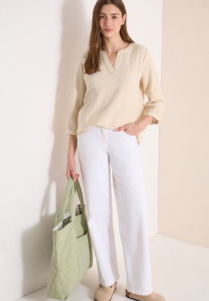 Kobieta ubrana w kremową bluzkę i białe spodnie trzyma dużą, jasnozieloną pikowaną torbę na ramię, stojąc na tle beżowej ściany.