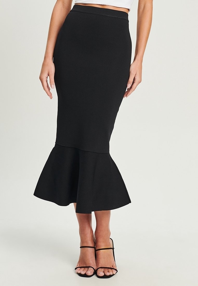 Femme portant une jupe midi noire style sirène associée à des sandales noires à talons hauts et brides, sur un fond uni.