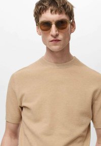 Pulover bej tricotat cu mânecă scurtă și guler rotund, asociat cu ochelari de soare cu tentă maronie, cu ramă aurie, textură netedă, design minimalist.