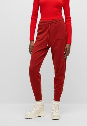 Persoon die rode gebreide joggerbroek met voorzakken draagt, een rood aangesloten top met lange mouwen en witte chunky veterlaarzen.