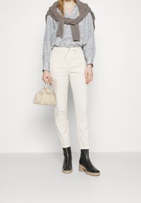 Vita bomullbyxor med dragsko i midjan, blåmönstrad blus, brunstickad tröja över axlarna, beige handväska, svarta klackboots.