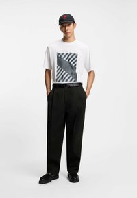 Maglietta bianca con stampa grafica nera, pantaloni neri a vita alta, cintura nera e scarpe nere. Il berretto è nero con logo rosso.