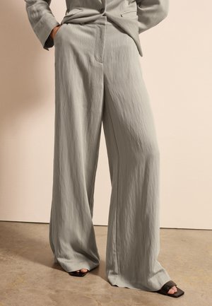 Helle graue weite Hose aus weichem, strukturiertem Stoff. Mit hoher Taille und seitlichen Taschen. Kombiniert mit schwarzen offenen Schuhen.